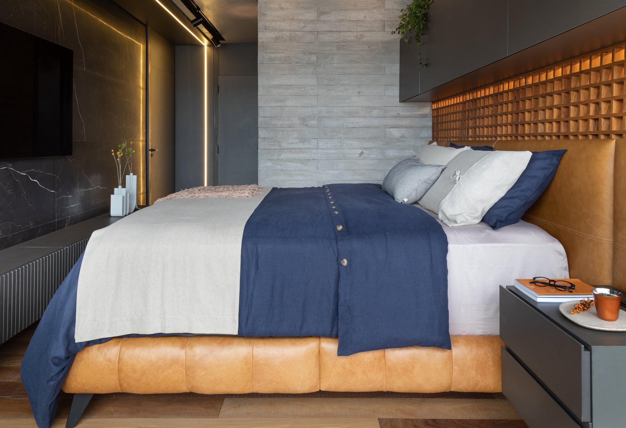 Projeto Mooca: Dormitório com cama de couro caramelo, painel de TV em mármore escuro e cabeceira ripada de madeira.
