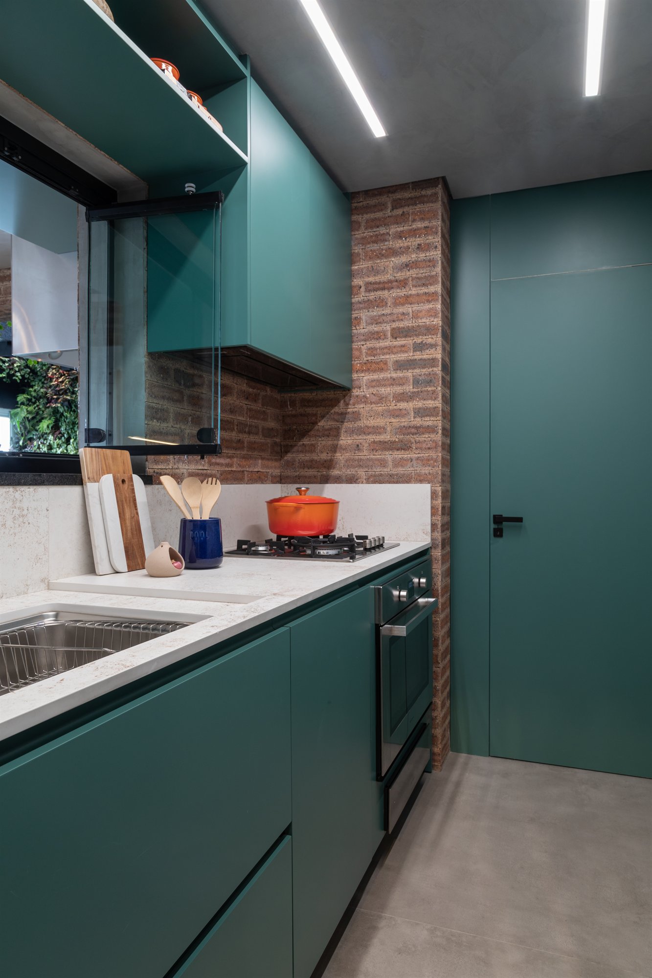 Projeto Mooca: Detalhe da cozinha verde com bancada, cuba, cooktop e parede de tijolos aparentes.