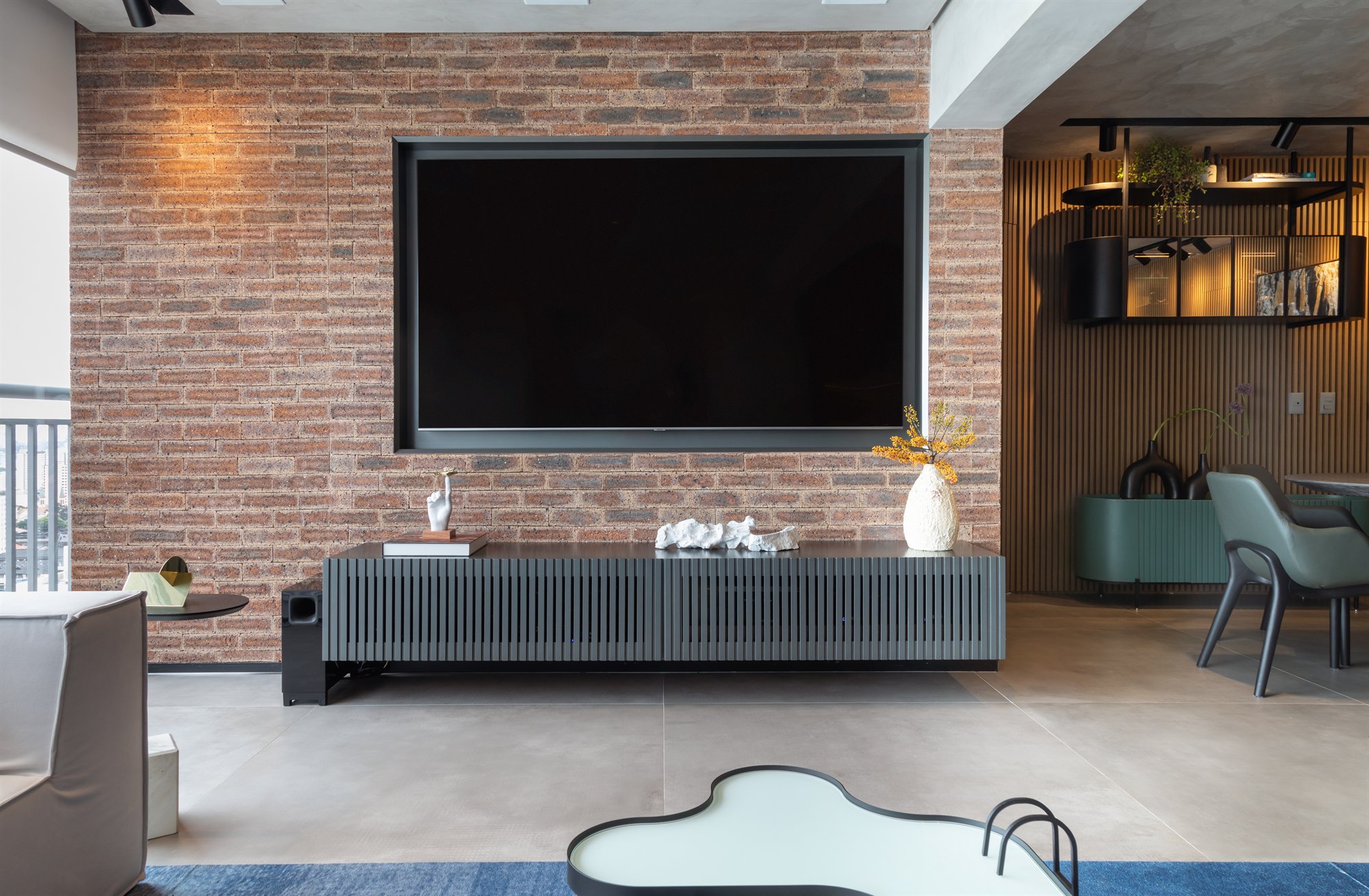Projeto Mooca: Parede de tijolos com TV e rack ripado, com espaço gourmet ao fundo.