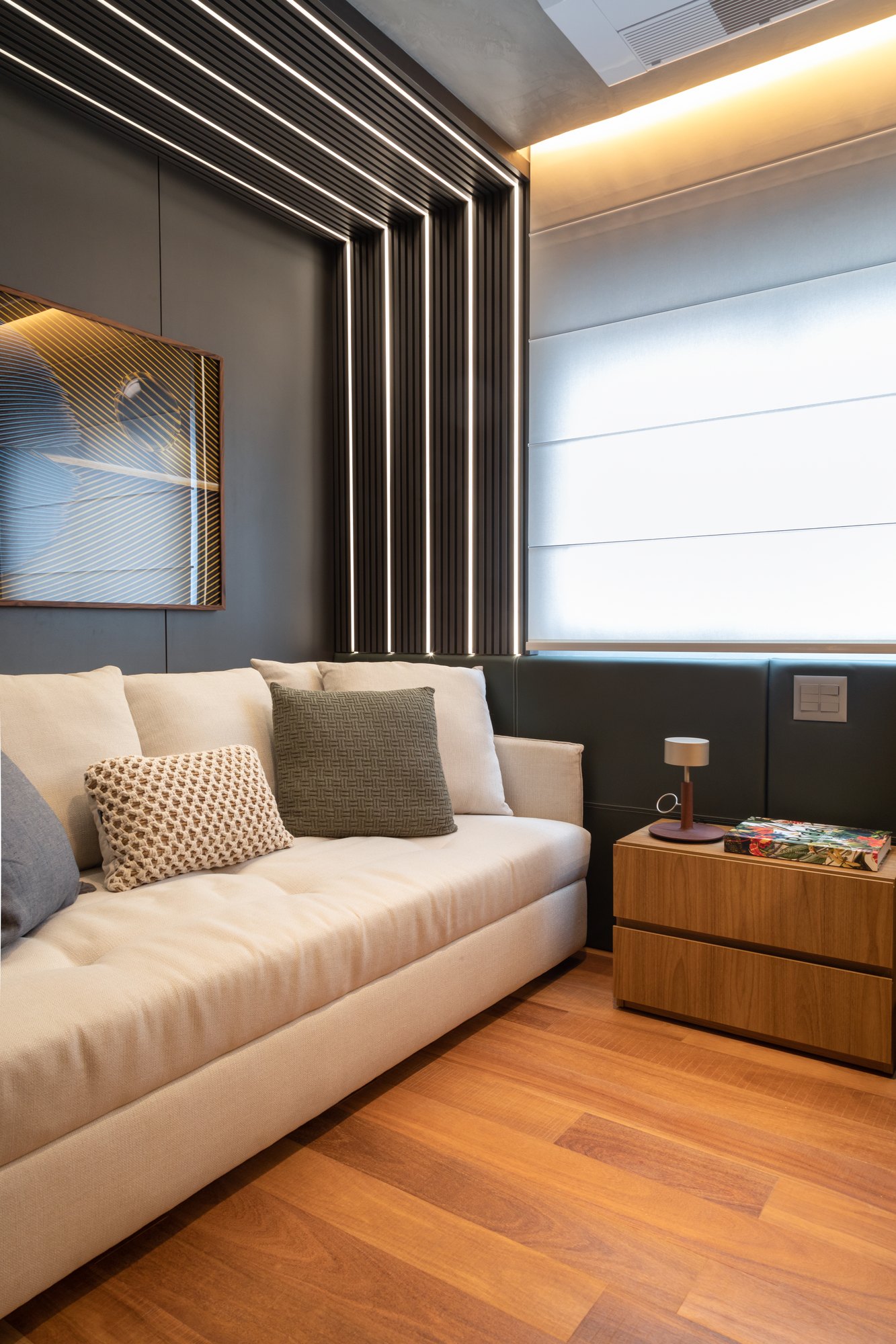 Projeto Mooca: Sofá claro junto a painel escuro com iluminação linear e mesa lateral de madeira.