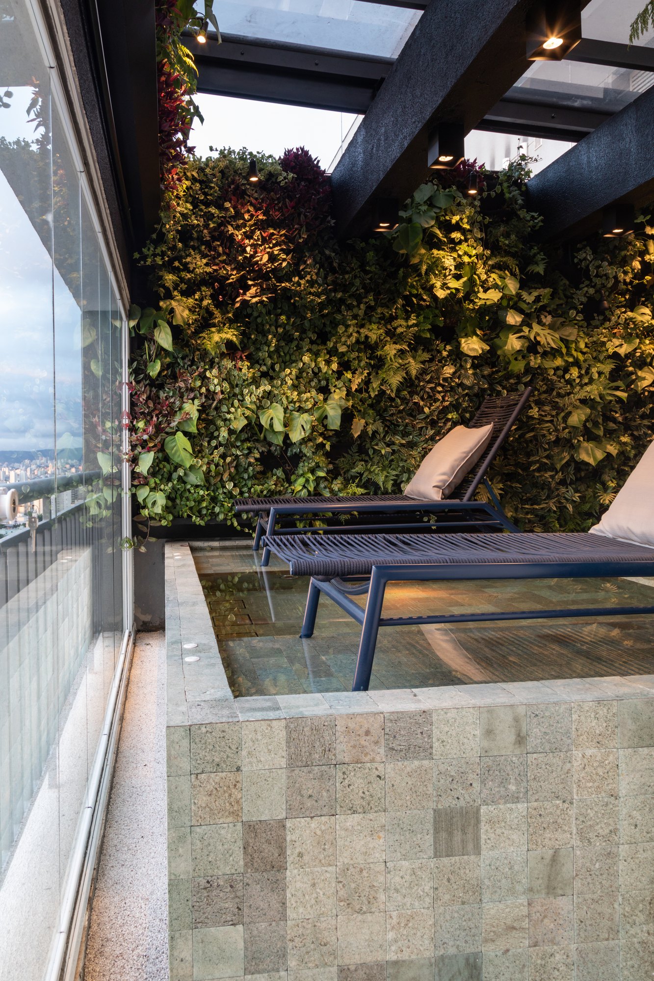 Projeto Mooca: Piscina na varanda junto a jardim vertical e espreguiçadeiras.