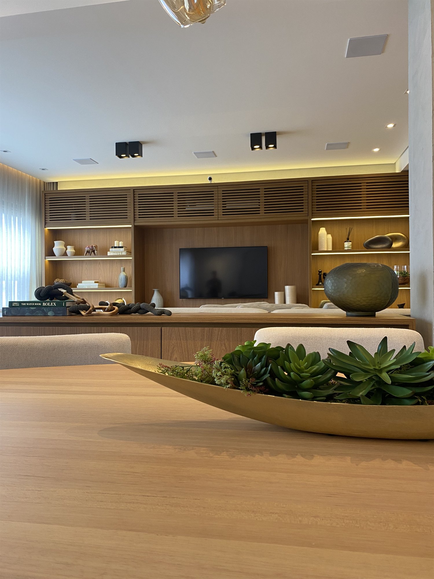 Projeto Itaim Bibi 2024: Mesa de jantar com arranjo decorativo diante do painel de TV em madeira.